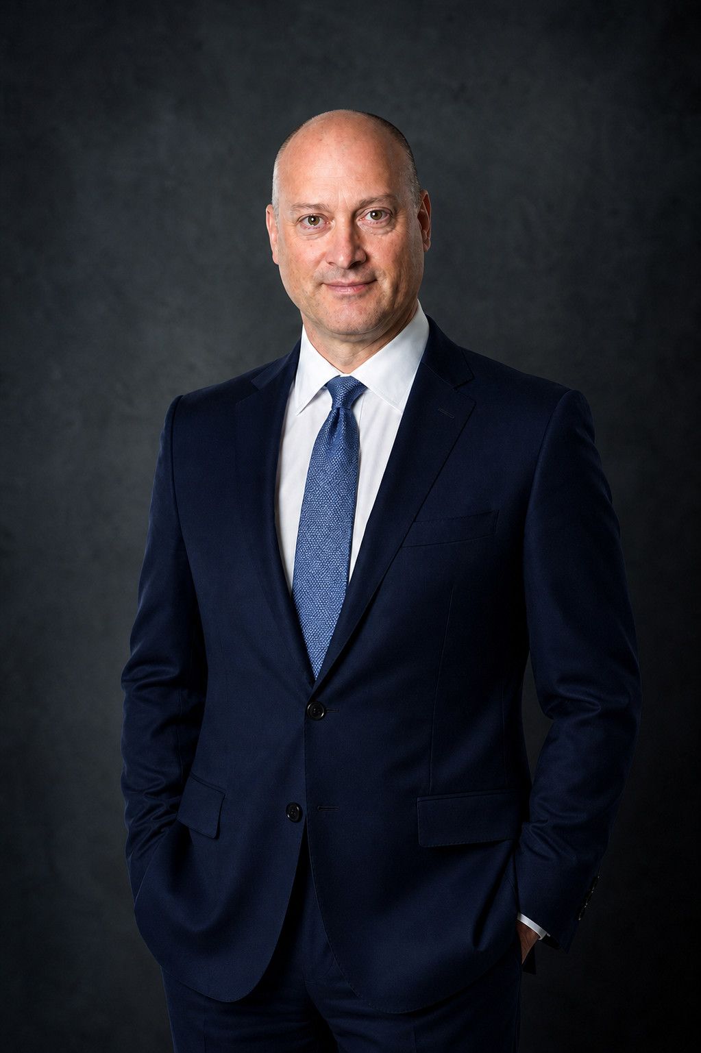 Peter DeCaprio in a navy suit and blue tie, standing against a dark studio backdrop
